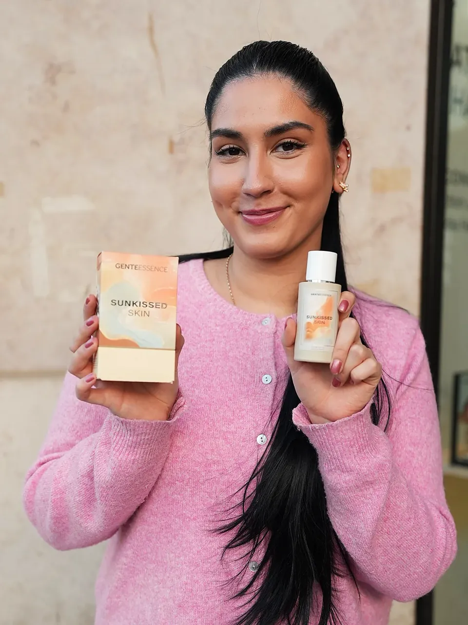 Sunkissed Skin: Eine Frau mit langem schwarzem Zopf und rosa Strickjacke hält Verpackung und Flakon. Originalfoto.